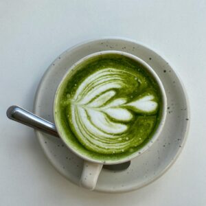 En kop matcha latte med latte art på toppen, serveret på en prikket tallerken med en ske